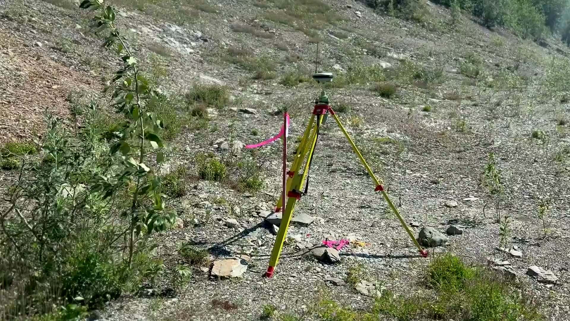 GNSS survey base station on a yellow tripod with flagging on a rocky mine site slope in summer
