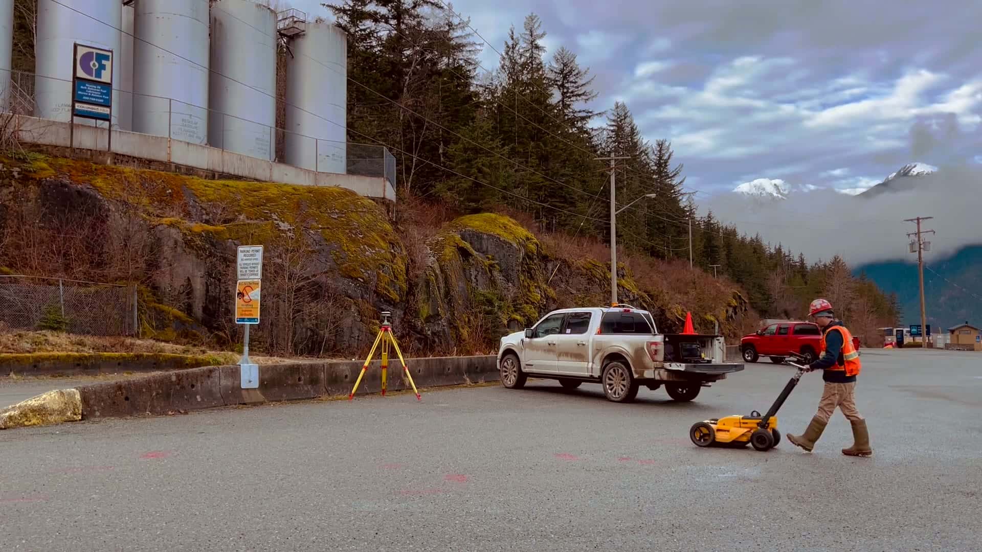 Technician pushing a ground penetrating radar cart across a paved lot below a rock bluff on the BC coast, with a GNSS tripod and survey truck beside him and snow peaks behind