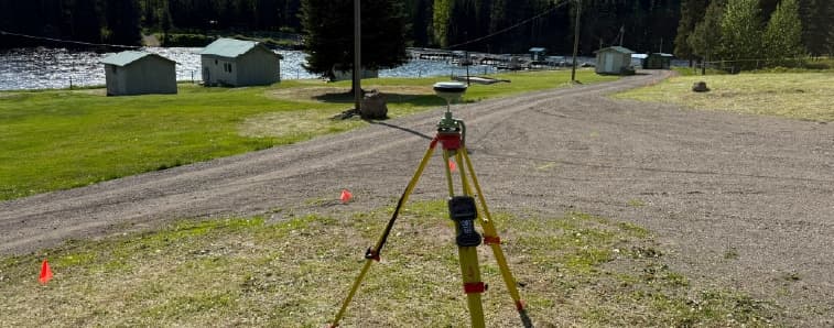 Surveying tripod with instrument set up on a lakeside path, with small sheds, cones, and boats in the background.
