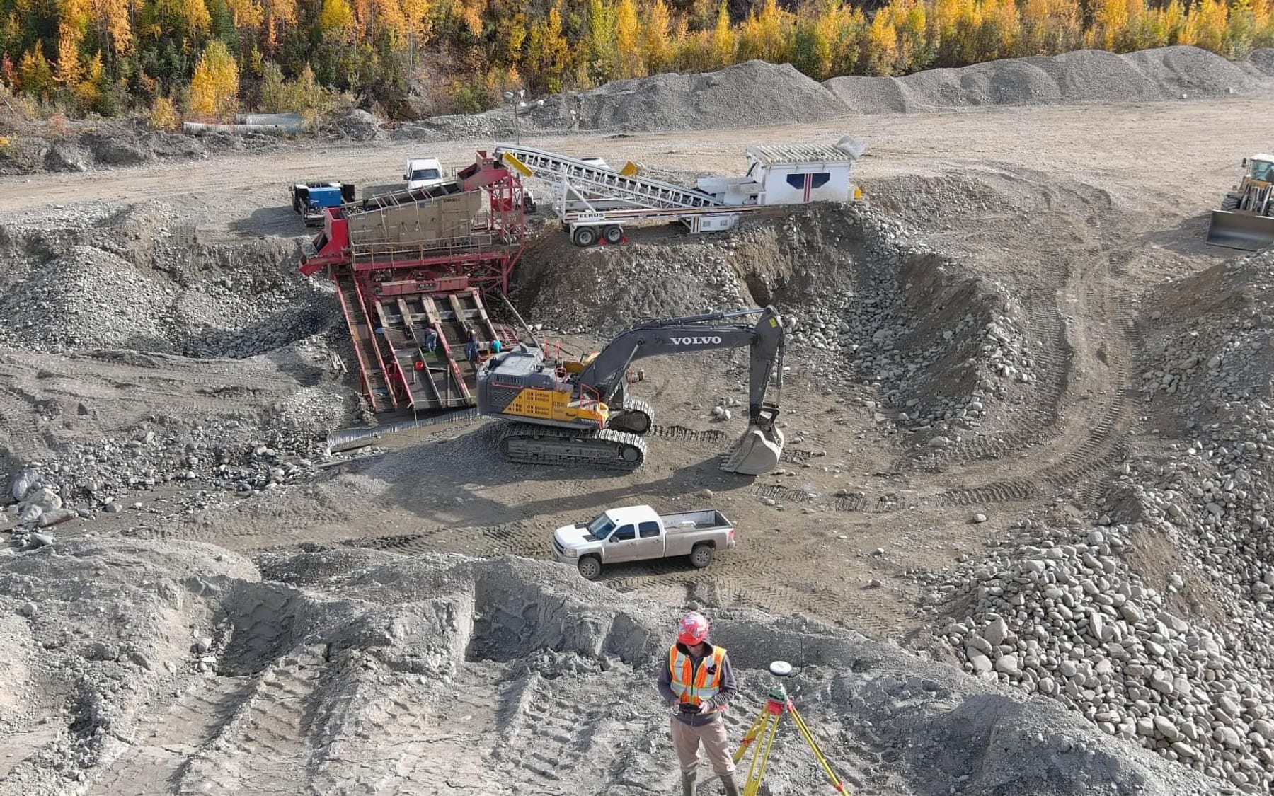 Aerial view of a placer gold operation in fall colours — wash plant and excavator running while a surveyor sets up a GNSS receiver in the foreground