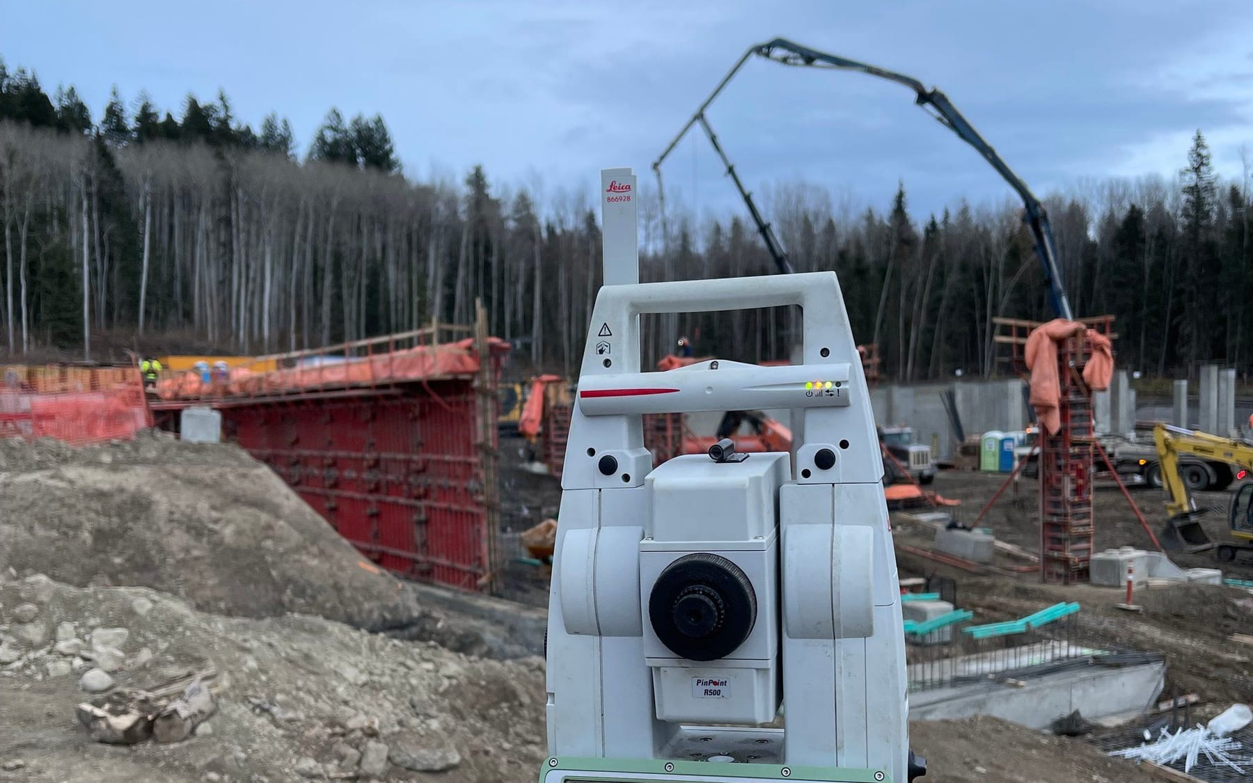 Robotic total station set up in front of formwork with a concrete pump boom overhead on an active construction site