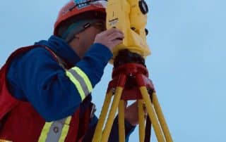 PCI Surveyor in winter gear using a total station on a tripod in a snowy landscape with trees and mountains in the background.