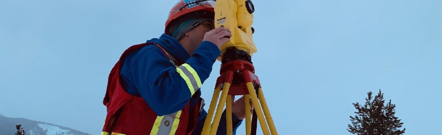 PCI Surveyor in winter gear using a total station on a tripod in a snowy landscape with trees and mountains in the background.