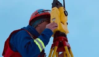 PCI Surveyor in winter gear using a total station on a tripod in a snowy landscape with trees and mountains in the background.