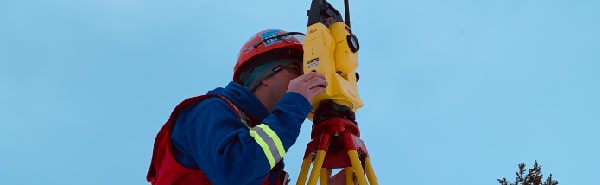 PCI Surveyor in winter gear using a total station on a tripod in a snowy landscape with trees and mountains in the background.