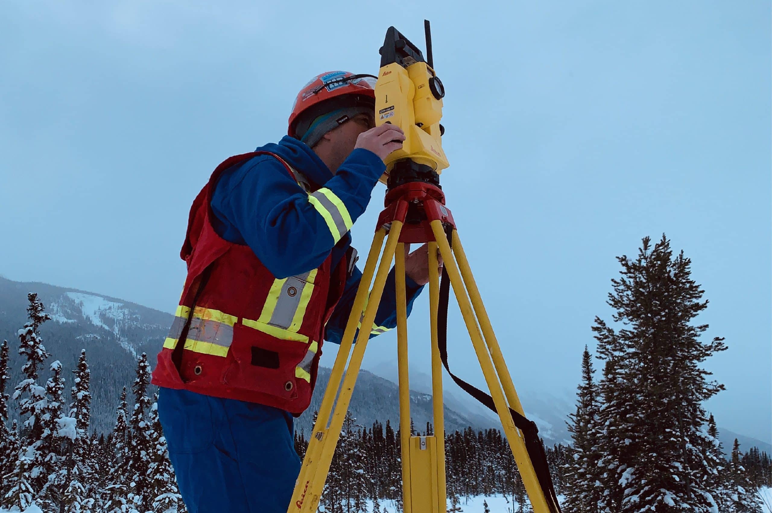 PCI Surveyor in winter gear using a total station on a tripod in a snowy landscape with trees and mountains in the background.