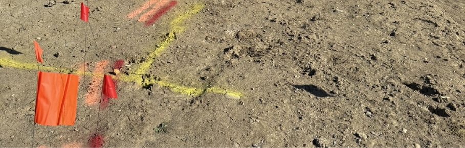 Utility locating markings and coloured flags on a construction site for drilling and excavation showing red, yellow, and orange lines and markers. Yellow is gas, orange is telephone and red is power/electric.
