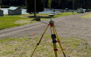 Surveying tripod with instrument set up on a lakeside path, with small sheds, cones, and boats in the background.