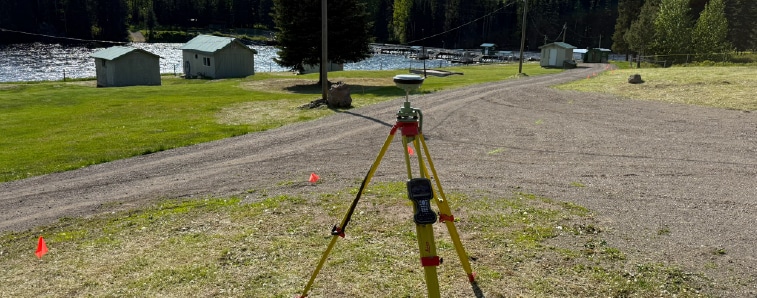 Surveying tripod with instrument set up on a lakeside path, with small sheds, cones, and boats in the background.